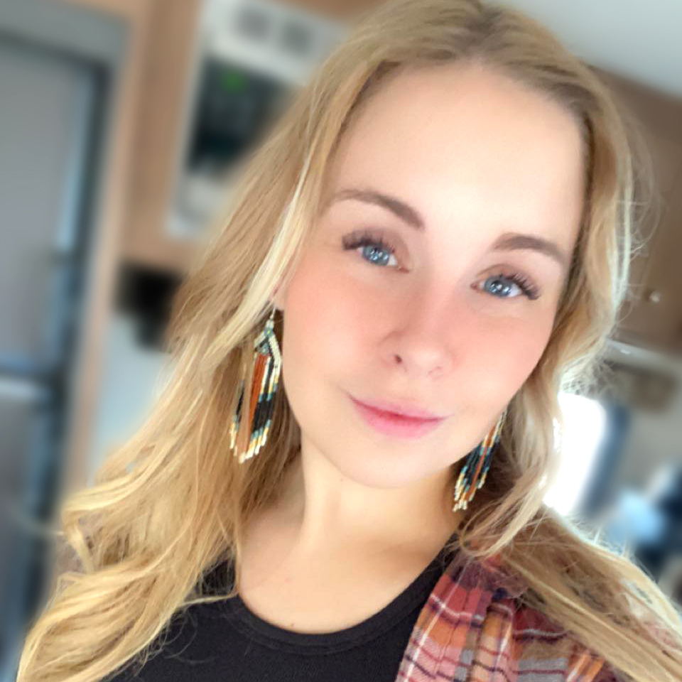 A young woman with long blonde hair wears dangling beaded earrings and a plaid shirt, smiling slightly while looking at the camera indoors.